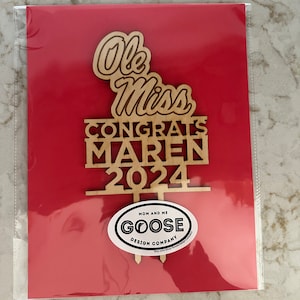 Puede incluir: Topper de pastel de madera con el texto "Ole Miss Congrats Maren 2024" y una pequeña pegatina ovalada que dice "Mom and Me Goose Design Company".