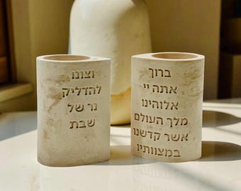 May include: Two off-white, cylindrical candle holders with Hebrew text. The holders have a textured surface and a rounded top. A large, off-white vase is in the background. The candle holders are on a light-colored surface.