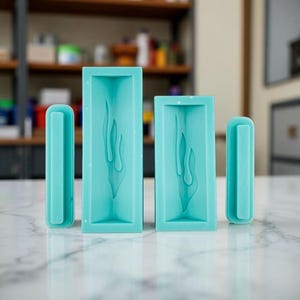 May include: Four teal silicone molds for crafting. Two rectangular molds feature a flame design, and two smaller, curved molds are also included. The molds are arranged on a white marble surface, with a blurred background of shelves and objects.