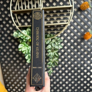 K&ouml;nnte beinhalten: Ein schwarzes, ledergebundenes Buch mit goldener Beschriftung auf dem R&uuml;cken, die "Chronik des Glases" von Sarah J. Maas lautet.