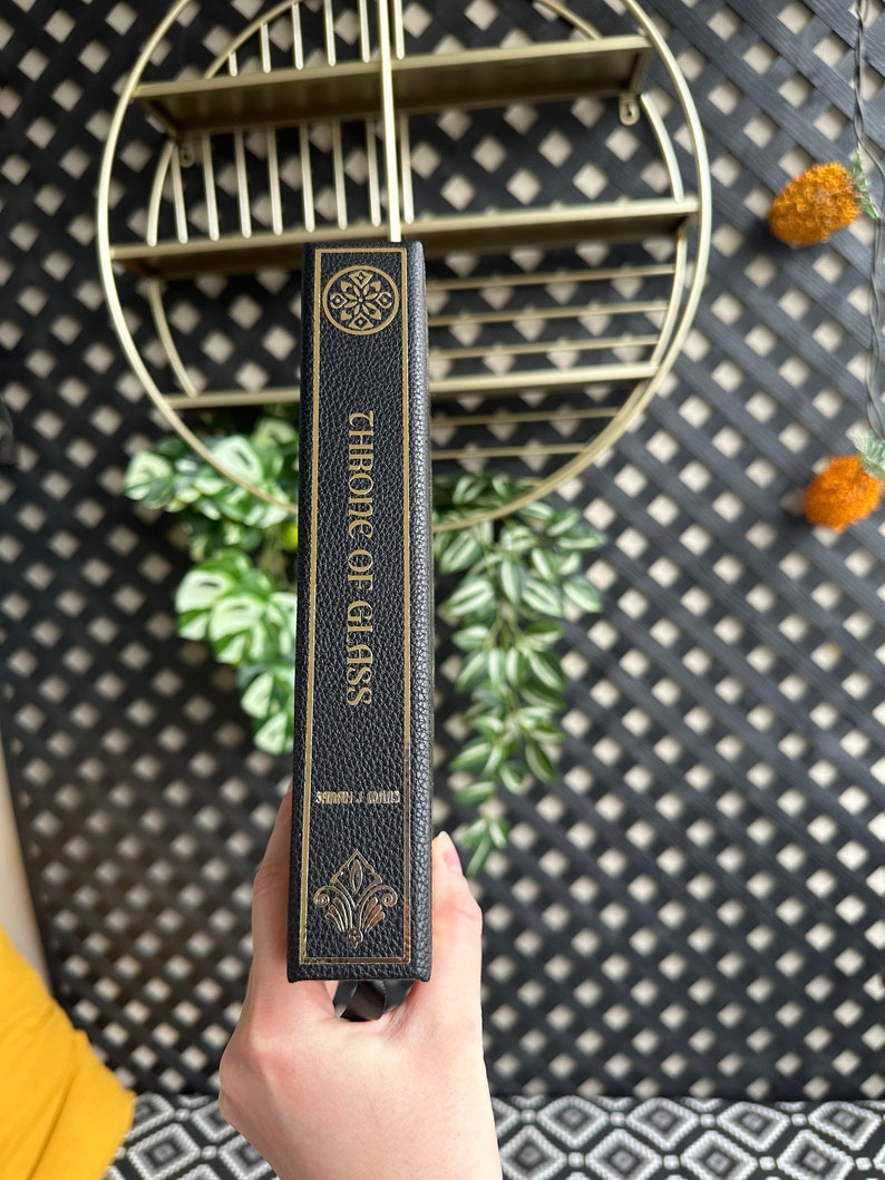 K&ouml;nnte beinhalten: Ein schwarzes, ledergebundenes Buch mit goldener Beschriftung auf dem R&uuml;cken, die "Chronik des Glases" von Sarah J. Maas lautet.