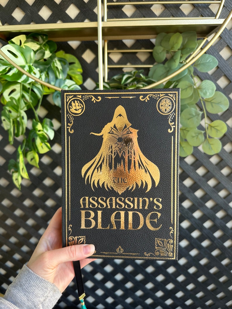 Op de afbeelding: Een zwart lederen gebonden boek met gouden letters en een gouden illustratie van een figuur in een kap. De titel van het boek is "The Assassin's Blade".