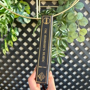 Op de afbeelding: Een zwart lederen gebonden boek met gouden letters. De titel is "The Assassin's Blade" door Jennifer L. Armentrout.