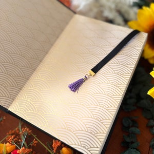 May include: Open black journal with a purple tassel bookmark. The journal has a white cover with a gold wave pattern.