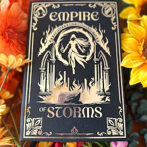 May include: A black leather-bound journal with gold lettering. The title is "Empire of Storms". The cover features a gold illustration of a woman in a flowing gown standing in a fiery archway.