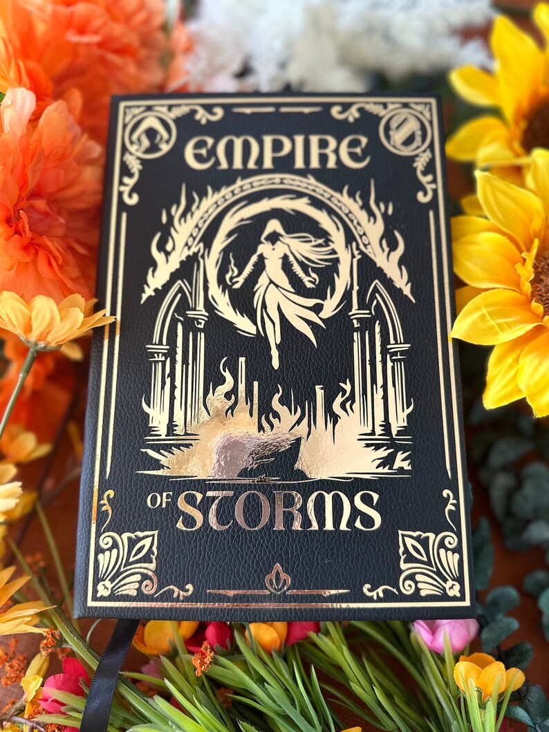 May include: A black leather-bound journal with gold lettering. The title is "Empire of Storms". The cover features a gold illustration of a woman in a flowing gown standing in a fiery archway.