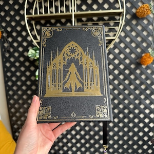 K&ouml;nnte beinhalten: Ein schwarzes, ledergebundenes Tagebuch mit einem goldgepr&auml;gten Design, das eine gotische Kathedrale und eine Frau im Eingangsbereich zeigt. Das Tagebuch hat ein schwarzes Band-Lesezeichen mit einem violetten Quasten.