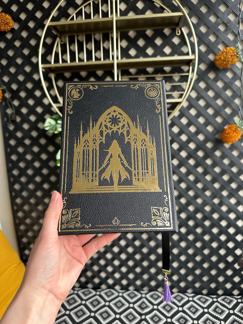 K&ouml;nnte beinhalten: Ein schwarzes, ledergebundenes Tagebuch mit einem goldgepr&auml;gten Design, das eine gotische Kathedrale und eine Frau im Eingangsbereich zeigt. Das Tagebuch hat ein schwarzes Band-Lesezeichen mit einem violetten Quasten.