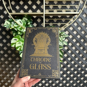 K&ouml;nnte beinhalten: Ein schwarzes Buch mit einem goldenen Pr&auml;gedesign eines Throns und den Worten "THRONE OF GLASS" auf dem Einband.