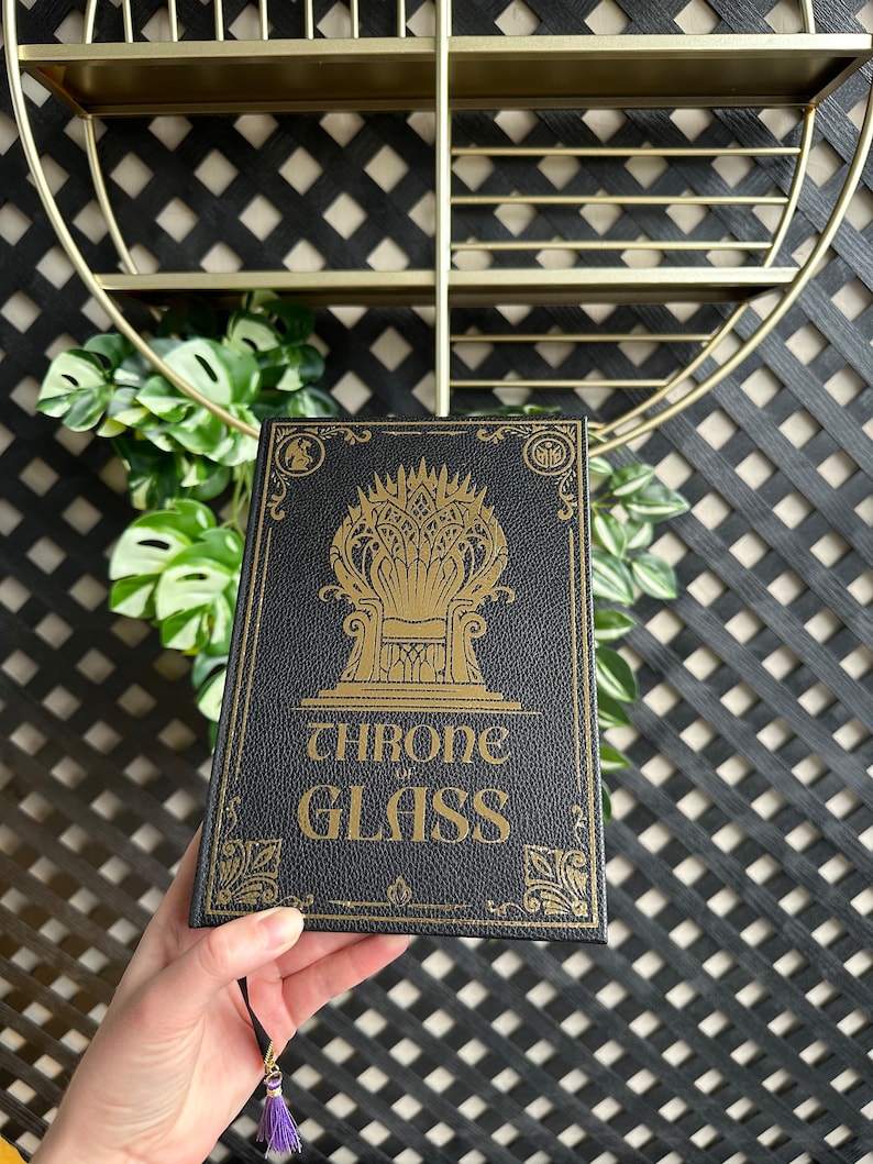 K&ouml;nnte beinhalten: Ein schwarzes Buch mit einem goldenen Pr&auml;gedesign eines Throns und den Worten "THRONE OF GLASS" auf dem Einband.