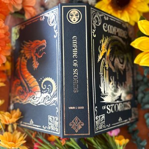 May include: A black leather-bound book with gold accents and a dragon design on the front cover. The title "Empire of Storms" is written in gold on the spine. The book is surrounded by yellow flowers.
