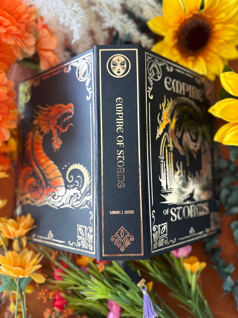 May include: A black leather-bound book with gold accents and a dragon design on the front cover. The title "Empire of Storms" is written in gold on the spine. The book is surrounded by yellow flowers.