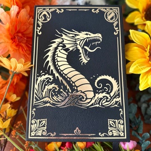 May include: Black leather journal with a gold embossed dragon design. The dragon is depicted with its mouth open and its tail curled around its body. The journal has a gold border and a gold ribbon bookmark.