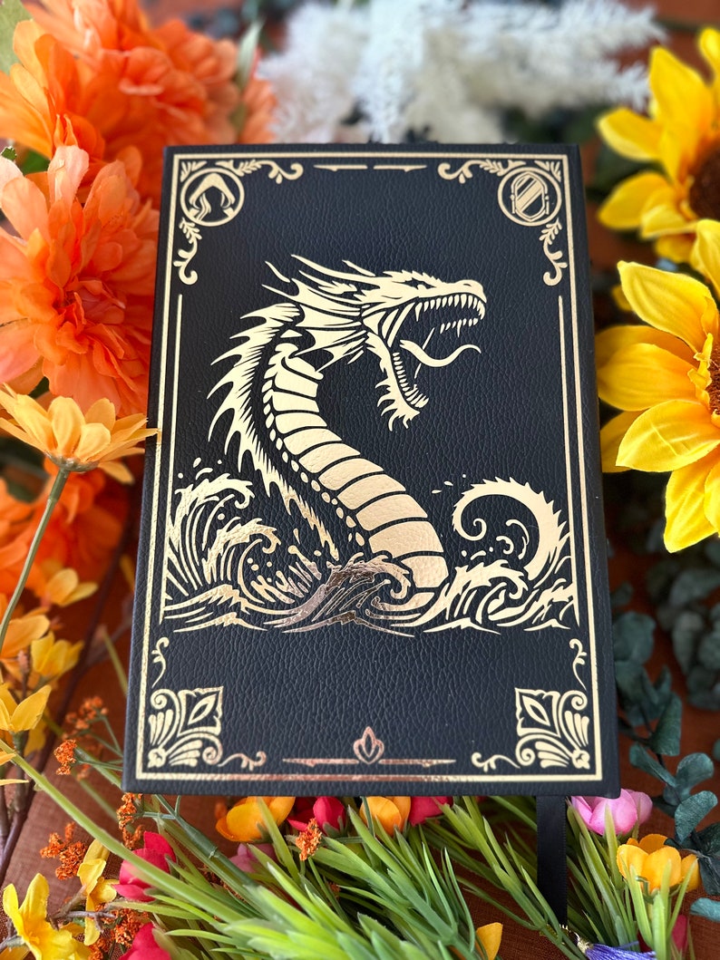 May include: Black leather journal with a gold embossed dragon design. The dragon is depicted with its mouth open and its tail curled around its body. The journal has a gold border and a gold ribbon bookmark.