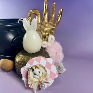 Op de afbeelding: Een grillige opstelling met een zwarte ketel, een gouden handsculptuur en diverse decoraties met een konijnenthema. Inclusief een witte konijnvormige kaars, een roze pluizig konijn en een decoratieve konijnenbroche met een strik en parel.