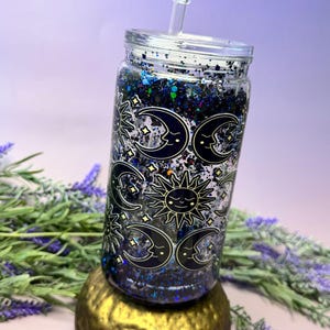 May include: A clear glass tumbler with a lid and straw, filled with dark blue and black glitter liquid. The exterior features a repeating pattern of black and gold sun and crescent moon faces with closed eyes, surrounded by small gold star shapes.