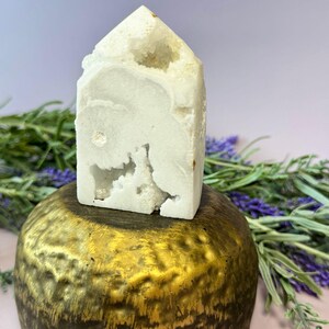 Op de afbeelding: Een witte kristallen toren met een getextureerd oppervlak en een puntige top. Het kristal staat op een goudkleurig, rond object. Groene en paarse bladeren omringen de objecten, tegen een lichtpaarse achtergrond.
