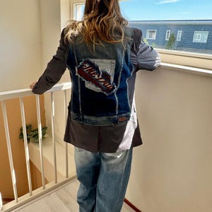 Op de afbeelding: Een spijkerjasje met een donkerblauw denim achterpaneel en grijze mouwen. De achterkant van de jas heeft een patch met de woorden "The Day" in rood en wit. De persoon op de foto draagt een lichtblauwe jeans.
