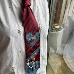 Op de afbeelding: Een bordeauxrode gestreepte stropdas met een denim hart en lippen applicatie. De stropdas heeft een denim zakje met een veiligheidsspeld en sieraden accenten. De stropdas is geknoopt en wordt gedragen over een wit overhemd met parels.