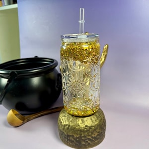 Op de afbeelding: Een doorzichtige glazen beker met een gouden glitterbasis en een deksel met een rietje. De beker is versierd met zon- en maanontwerpen. Hij staat op een goudkleurige standaard, naast een zwarte ketel en een houten lepel.