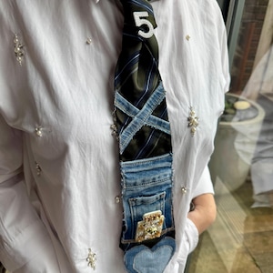 Op de afbeelding: Een unieke stropdas met een zwart-groen gestreept patroon, geaccentueerd met denim patches en een denim zak. Een strass nummer 5 is aan de stropdas bevestigd. De stropdas heeft een hartvormige denim punt.