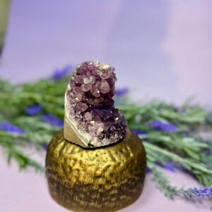 Op de afbeelding: Een cluster van dieppaarse amethistkristallen staat op een getextureerde, goudkleurige metalen basis. De basis heeft een afgeronde vorm. De achtergrond is zachtpaars met lavendel takjes. De kristallen zijn kristalachtig.