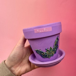 Può includere: Un vaso di fiori dipinto di viola con un piattino abbinato. Il vaso è decorato con un disegno di pipistrello realizzato con perline iridescenti. Un adesivo con la scritta "€6.50" è sul vaso.