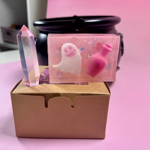 Op de afbeelding: Een roze kristal, een roze zeepblok met een spook- en drankontwerp en een kleine bruine doos. De zeepblok heeft een spook- en drankontwerp. Een zwarte ketel staat op de achtergrond. De items staan op een roze ondergrond.