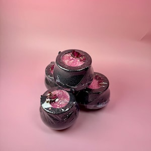 May include: A stack of four black cauldron-shaped bath bombs, each filled with pink granules and topped with dried rose petals. The bath bombs are wrapped in clear plastic, set against a pink background.