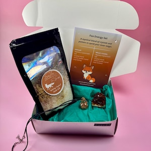 Op de afbeelding: Een open witte doos met een turquoise interieur bevat een zwarte zak met badzout, een kaart met een vosafbeelding, een kristal en een ketting. De kaart leest "Fox Energy Set" en geeft de inhoud weer. De doos staat op een roze ondergrond.