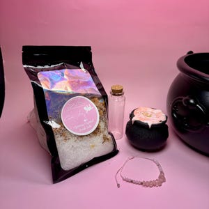 May include: A selection of self-care products on a pink surface. The image features a black bag of bath salts with a pink label, a small glass bottle, a black cauldron-shaped bath bomb, a rose quartz bracelet, and a black cauldron.