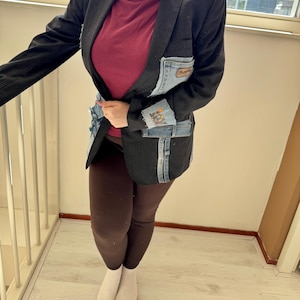 Op de afbeelding: Een zwarte krijtstreep blazer met denim patches en zakken. De blazer heeft een uniek ontwerp met denim accenten op de voorkant en mouwen. De zakken zijn voorzien van het Wrangler-logo. De blazer wordt gecombineerd met een bordeauxrode top en bruine leggings.