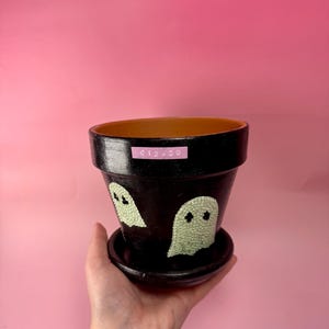 Op de afbeelding: Zwart geschilderde keramische bloempot met bijpassende schotel. De pot heeft twee lichtgroene spookontwerpen. De pot wordt in een hand gehouden tegen een roze achtergrond. De pot heeft een prijskaartje.
