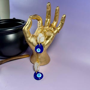 Op de afbeelding: Een goudkleurig handbeeld houdt een ketting vast met blauwe boze ooghangers en heldere kristallen stenen. Een zwarte ketel en een houten lepel staan op de achtergrond. De items staan op een paars oppervlak.