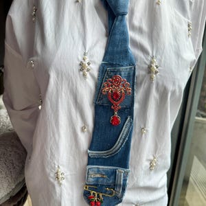 Op de afbeelding: Een unieke spijkerdas met een rode, met juwelen bezette versiering en een klein spijkerbroekzakje. De stropdas heeft een gouden veiligheidsspeld met rode kersenbedels. De stropdas wordt gedragen over een wit overhemd.