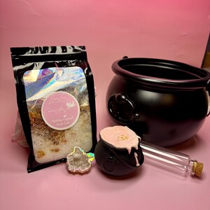 May include: A black cauldron, a small black cauldron with pink wax, a clear glass bottle with a cork, a geode, and a bag of bath salts are arranged on a pink surface. The bath salts are in a black bag with a holographic design.