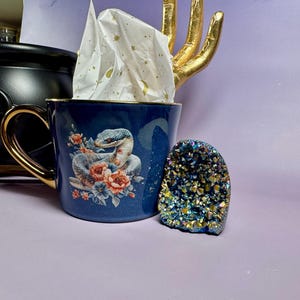 Pode incluir: Uma caneca de cerâmica azul escura com alça e borda douradas, com um design de cobra e floral. Uma geodo e uma estátua de mão dourada também são visíveis. A caneca está cheia de papel de seda.