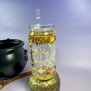 May include: A clear glass tumbler with a gold glitter and celestial design. The tumbler features a sun and moon design and comes with a clear straw and lid. A black cauldron and wooden ladle are in the background.