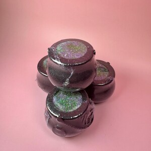 Può includere: Quattro bombe da bagno nere a forma di calderone con glitter iridescenti all'interno, avvolte in plastica trasparente. Le bombe da bagno sono disposte su uno sfondo rosa, creando un'esposizione stravagante e festosa.