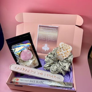 Op de afbeelding: Een roze doos met diverse zelfzorgartikelen. Het bevat badzout, een scrunchie, een keramische wierookhouder met maanfase-ontwerp en wierookstokjes. De doos bevat ook een kaart met de tekst "Sacred Calm & Cleansing Set".