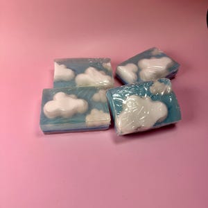 May include: Four rectangular soaps with a blue and white cloud design. The soaps are wrapped in clear plastic and arranged on a pink background. The soaps have a blue base with white cloud shapes.