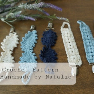 Può includere: Sei morsi per neonati all'uncinetto in bianco, blu e blu navy con clip argentati. I morsi sono realizzati con una varietà di punti e motivi all'uncinetto. "Crochet Pattern by Handmade by Natalie" è scritto su uno stendardo bianco.