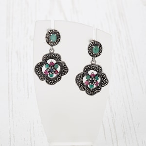 May include: Silver earrings with a floral design featuring green and pink gemstones. The earrings are on a white display stand.