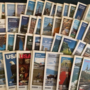 May include: A large collection of rectangular travel maps and guidebooks featuring various North American states and provinces. Each cover displays a unique regional photograph, bold text labels, and the AAA logo at the bottom of the white borders.