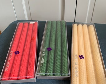May include: Three sets of honeycomb candles in red, green, and cream colors. Each set contains three candles and is packaged in a clear plastic box with the brand name "Simon Pearce" printed on the front.