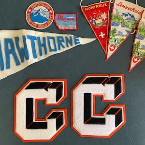 May include: A collection of vintage felt pennants and patches including a large white pennant with blue text reading HAWTHORNE, two triangular Lenzerheide pennants, a red Montreux pennant, a Switzerland patch, a Ski Bridger patch, and two large letter C patches.