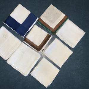 May include: An assortment of folded white handkerchiefs, some with decorative borders in navy blue, brown, and green. Several handkerchiefs feature an embroidered monogram. The items are arranged on a dark blue surface.