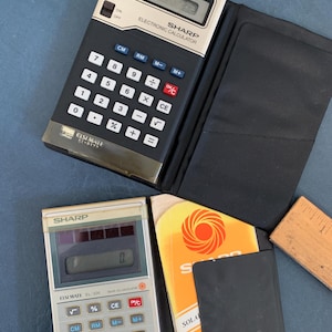 May include: Two vintage Sharp electronic calculators, one black and silver, the other silver and beige. The black calculator is open, revealing a display and buttons. A black case, a manual, and a wooden ruler are also present.