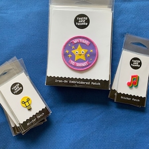 May include: Three iron-on patches with the text "These are things" and a lightbulb, a star with a face and the text "My time to shine!" and a musical note. The patches are packaged in clear plastic.