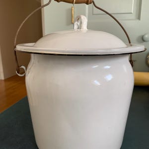 May include: A white enamel bucket with a matching lid and a dark metal rim. The bucket features a curved metal handle with a cylindrical wooden grip in the center. The surface is smooth and glossy, showing a simple, functional design.